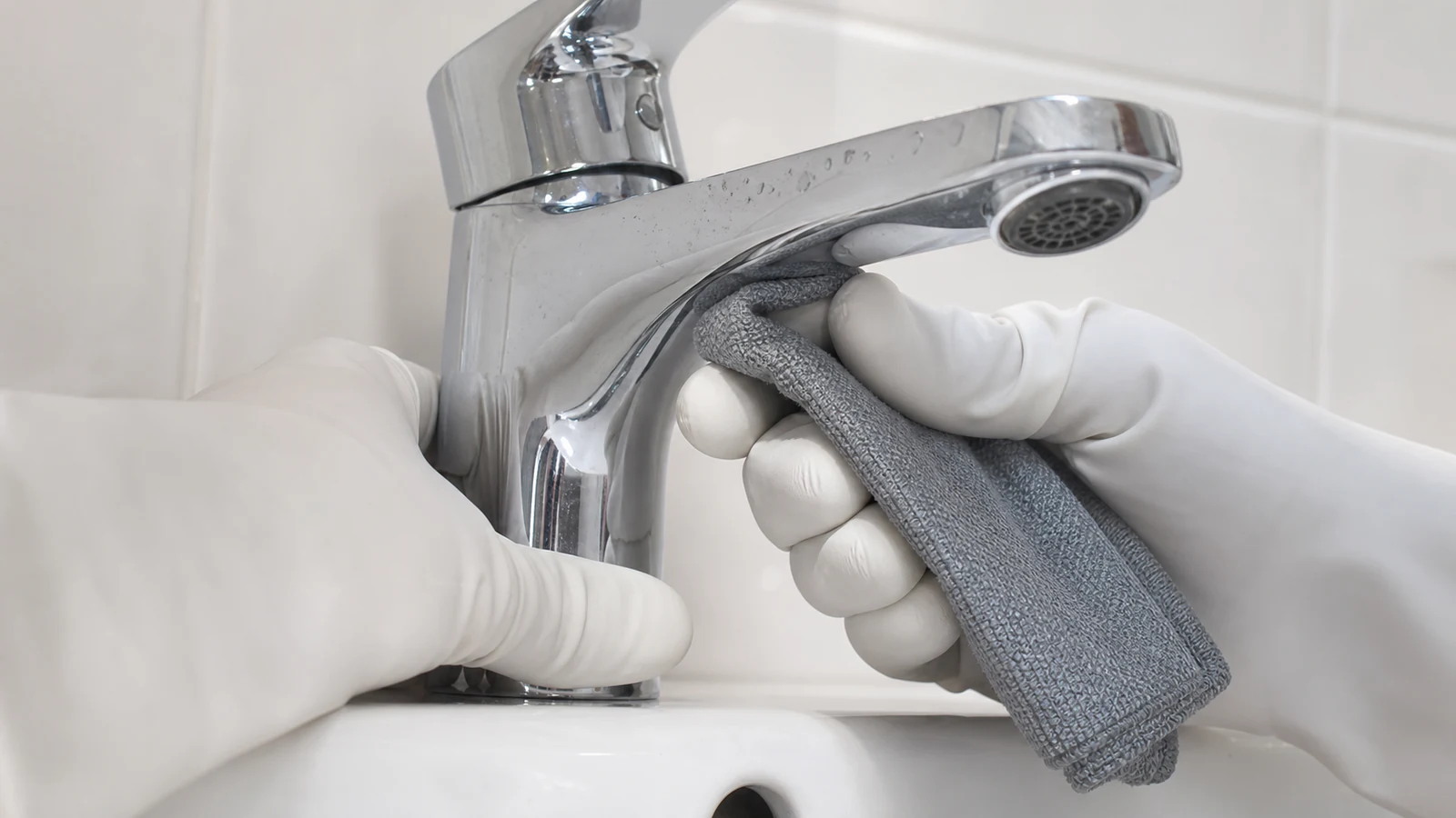 Gloved hand applying diluted cleaner to underside of tap spout with microfibre cloth