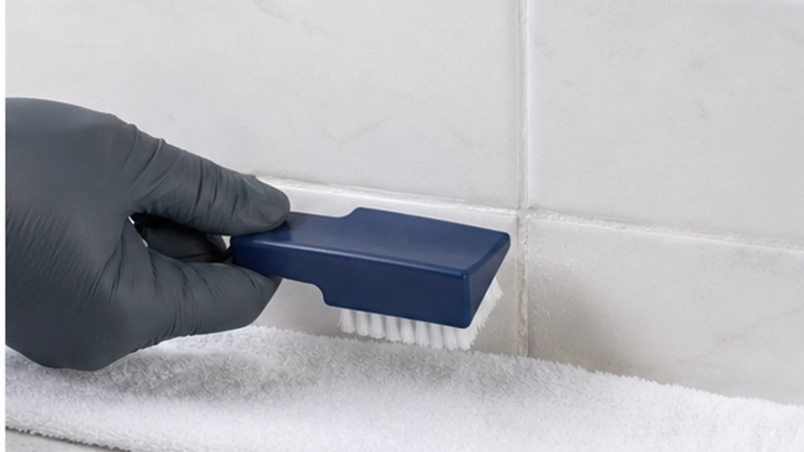 Gloved hand holding an old toothbrush, applying white paste to a dark grout line on backsplash tiles using small circular motions.
