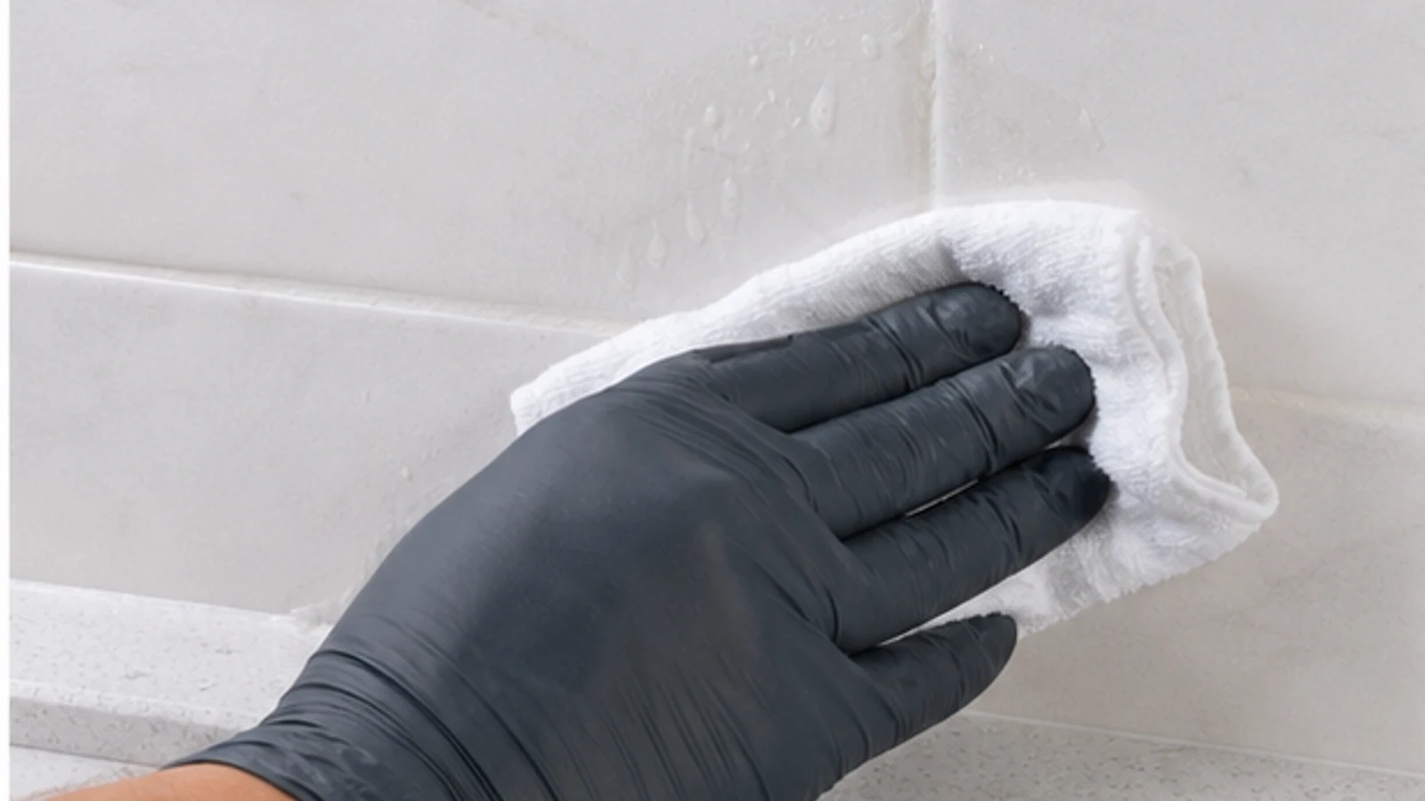 Gloved hand using a damp cloth to rinse a grout line after scrubbing, with white residue and water visible on the cloth.