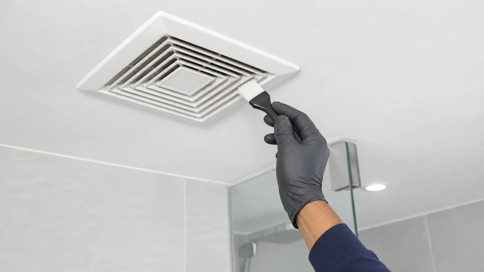 Overhead shot of a hand with a soft-bristled brush cleaning dust from a ceiling extractor fan grille, contemporary bathroom.