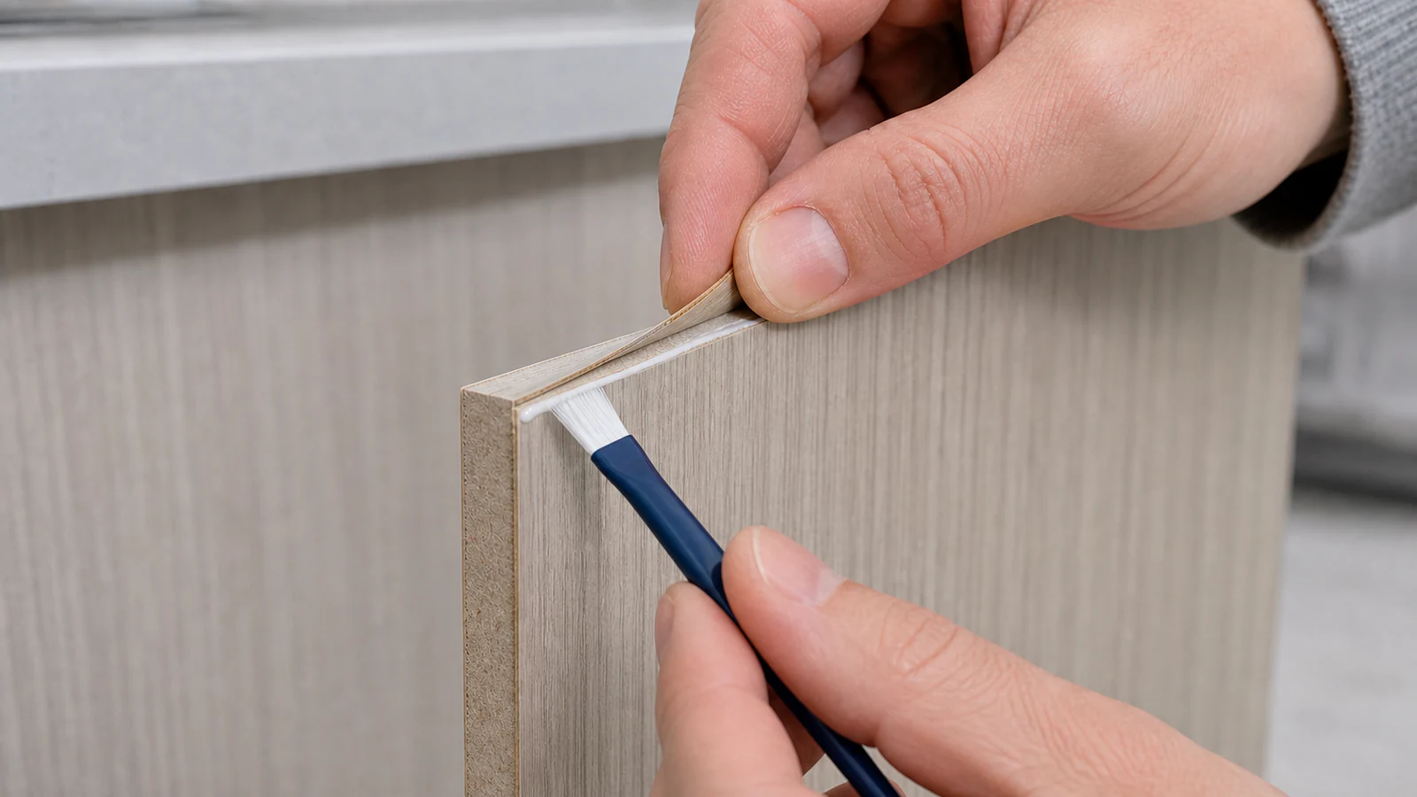 A small disposable brush applying a thin bead of wood glue along the substrate edge where the veneer will be pressed down, in neutral light.