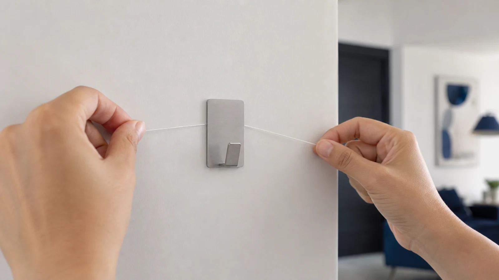 Dental floss positioned behind an adhesive hook on a painted wall, ready to slide gently downward to separate adhesive from surface.
