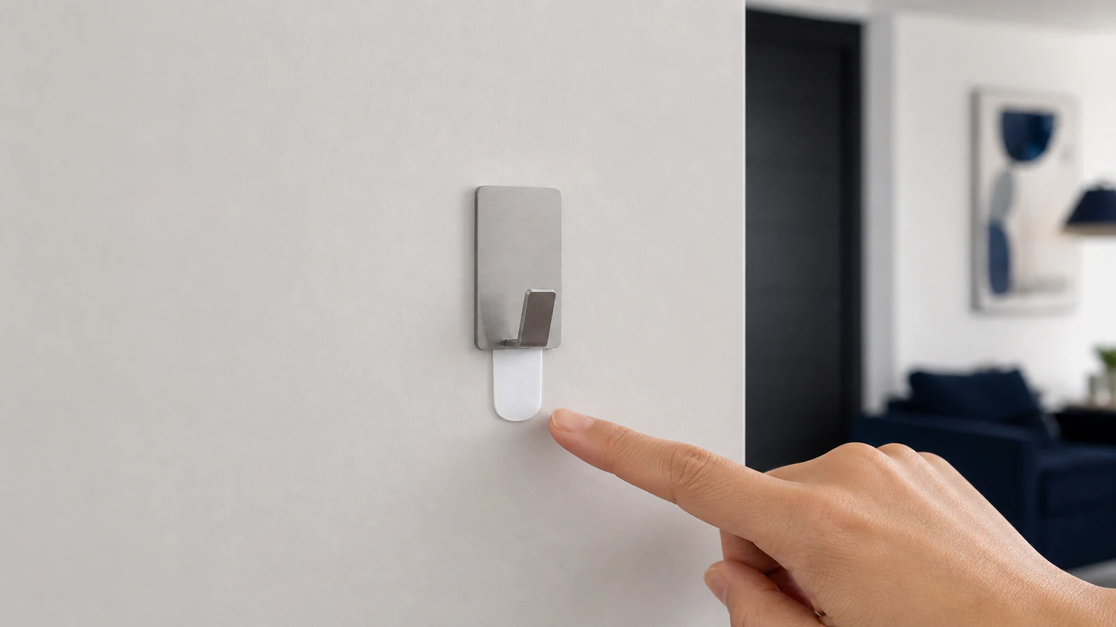 Hand pointing to the small white pull tab on the underside of an adhesive hook mounted on a painted wall, with the packet of hooks visible nearby showing printed instructions.