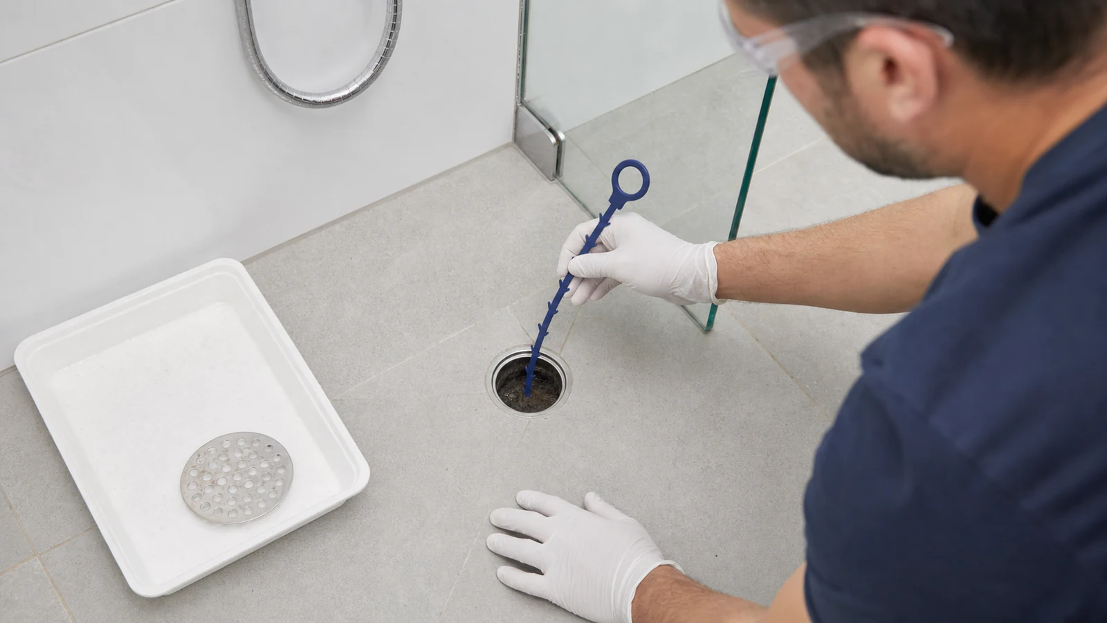 Gloved hand inserting a plastic drain hook into the drain opening at a shallow angle, with the hook edge visible.