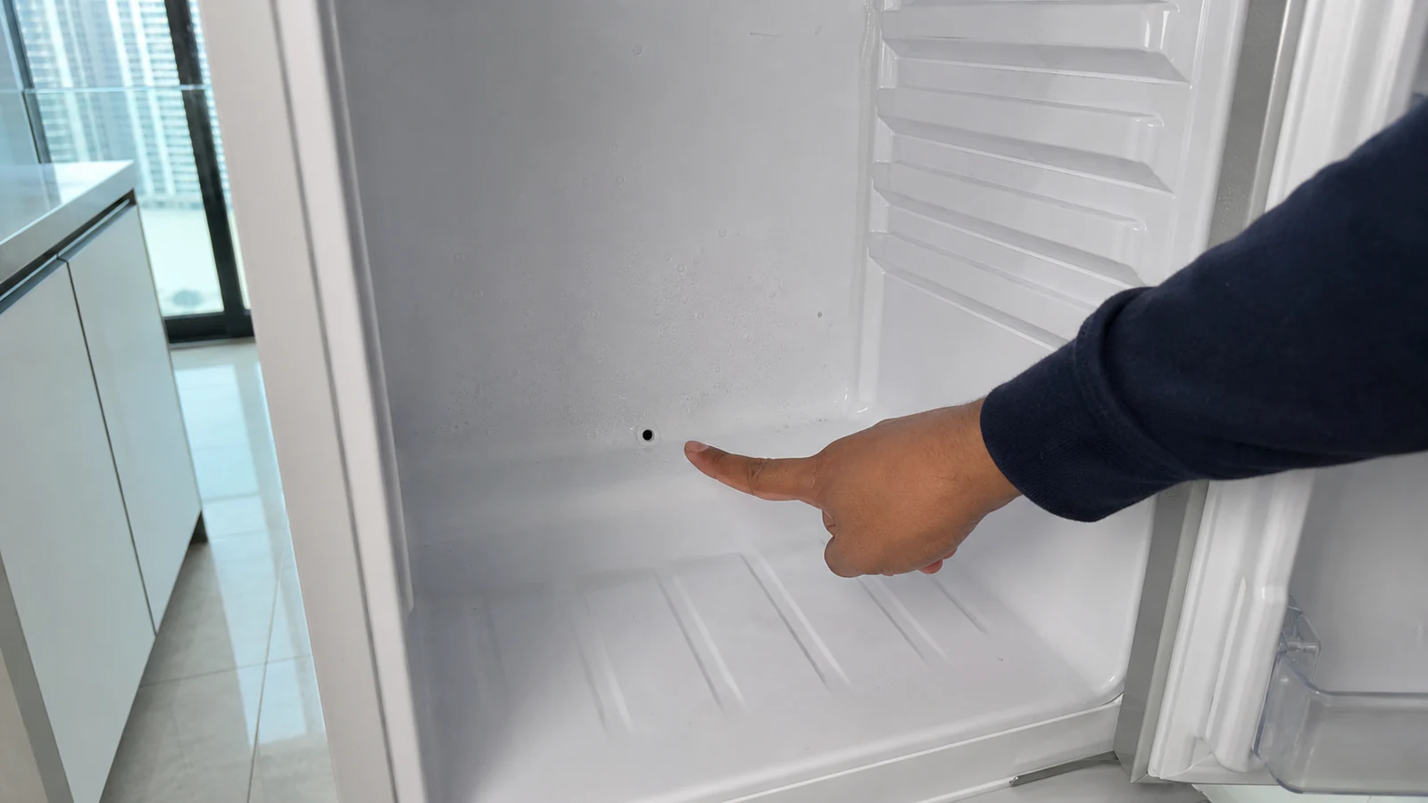 Fingertip pointing to a small drain hole visible at the lower rear interior of an empty refrigerator cabinet.