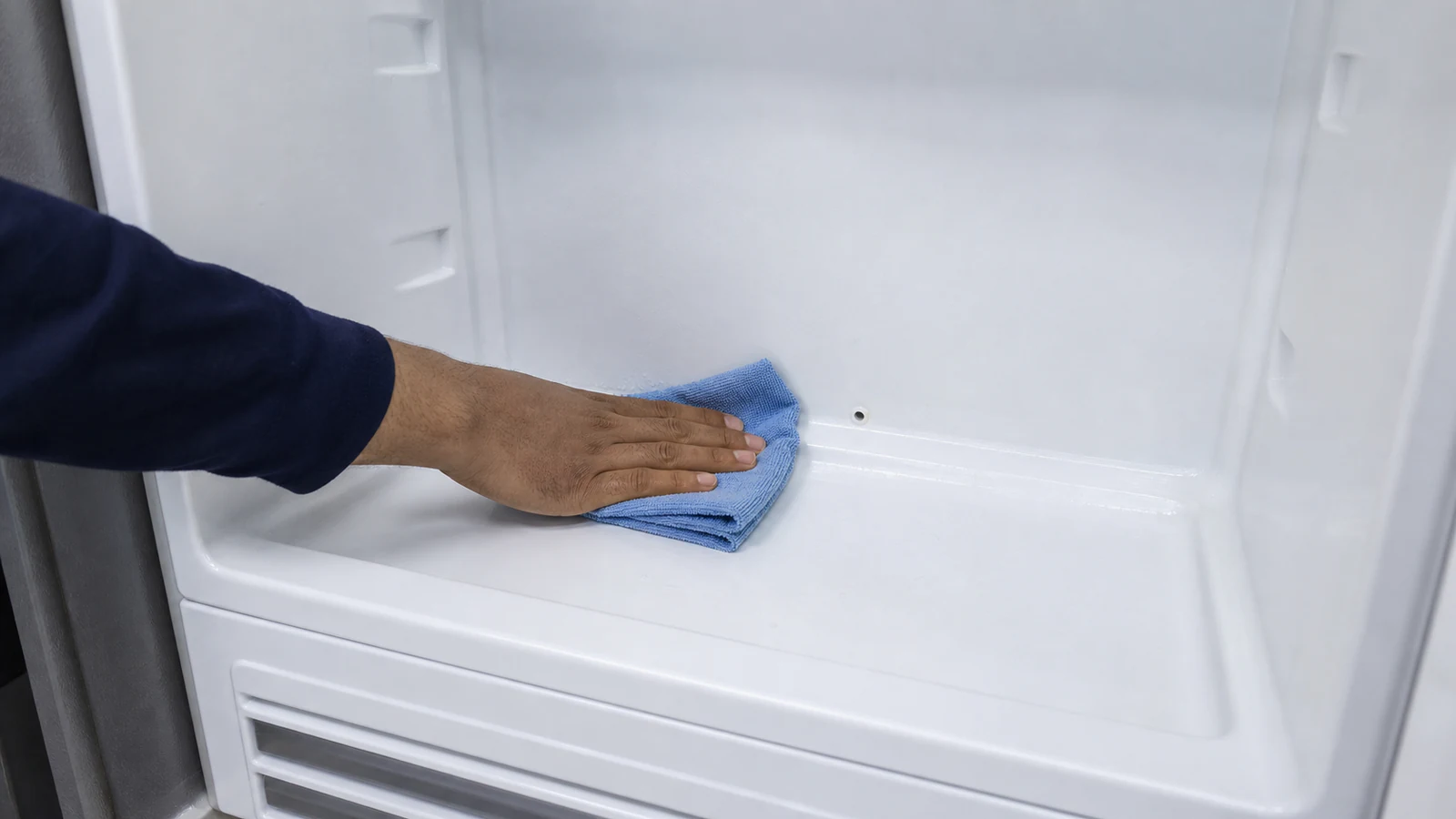 A damp microfibre cloth wiping the interior wall of an empty refrigerator with baking soda solution.