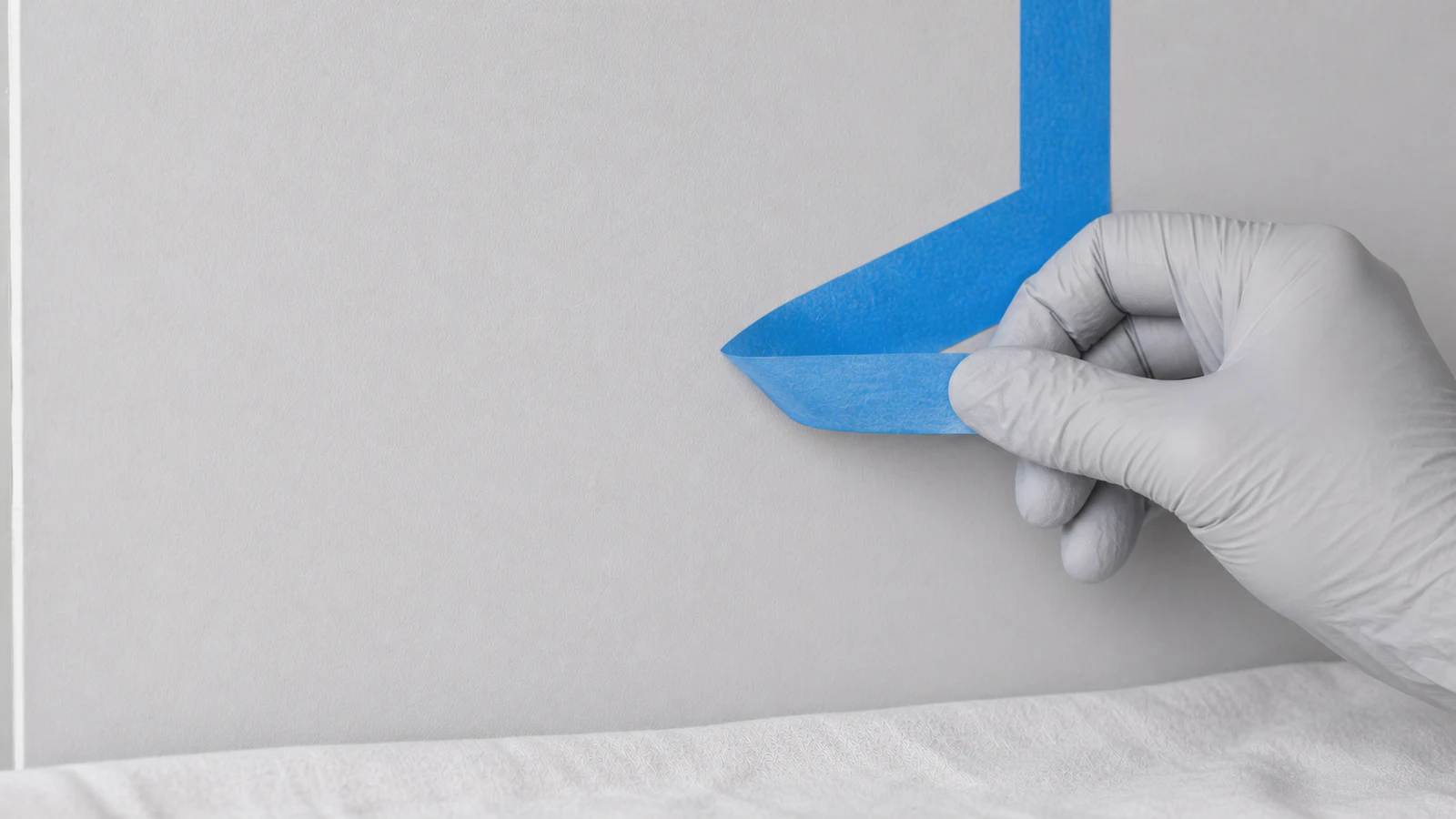 Gloved hand carefully peeling painter's tape away from a completed wall patch at a shallow angle, revealing a seamlessly blended painted area with no visible hard edge.