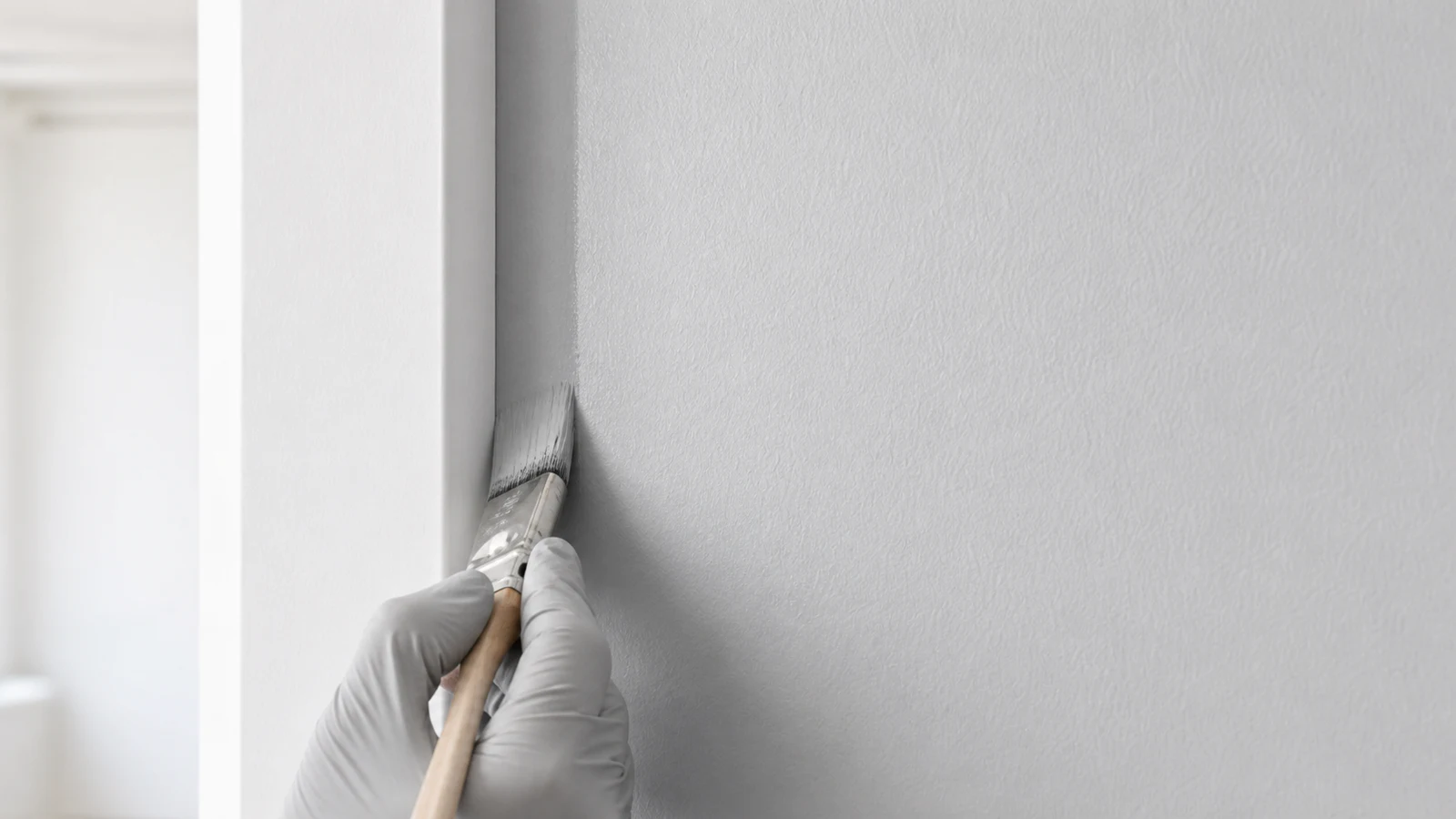 Gloved hand holding an angled brush, cutting a clean paint line from a wall patch to the corner, the brush edge sharp and precisely following the vertical corner line.