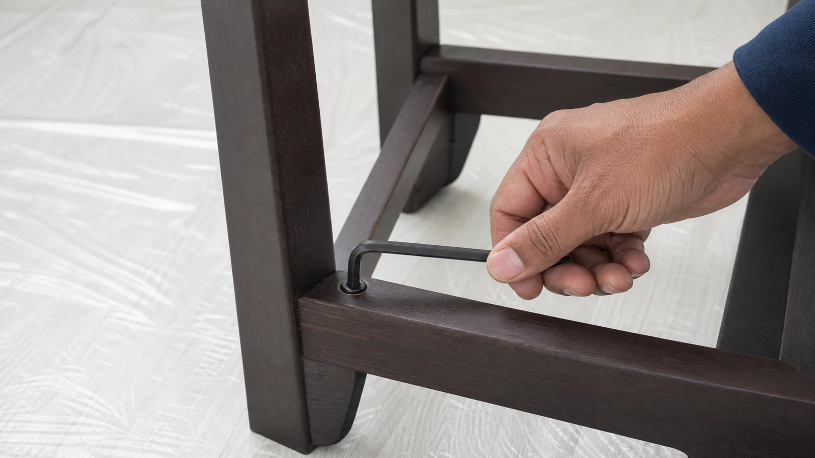 Hand turning an Allen key clockwise on an upturned chair, showing the fastener and connected leg frame.